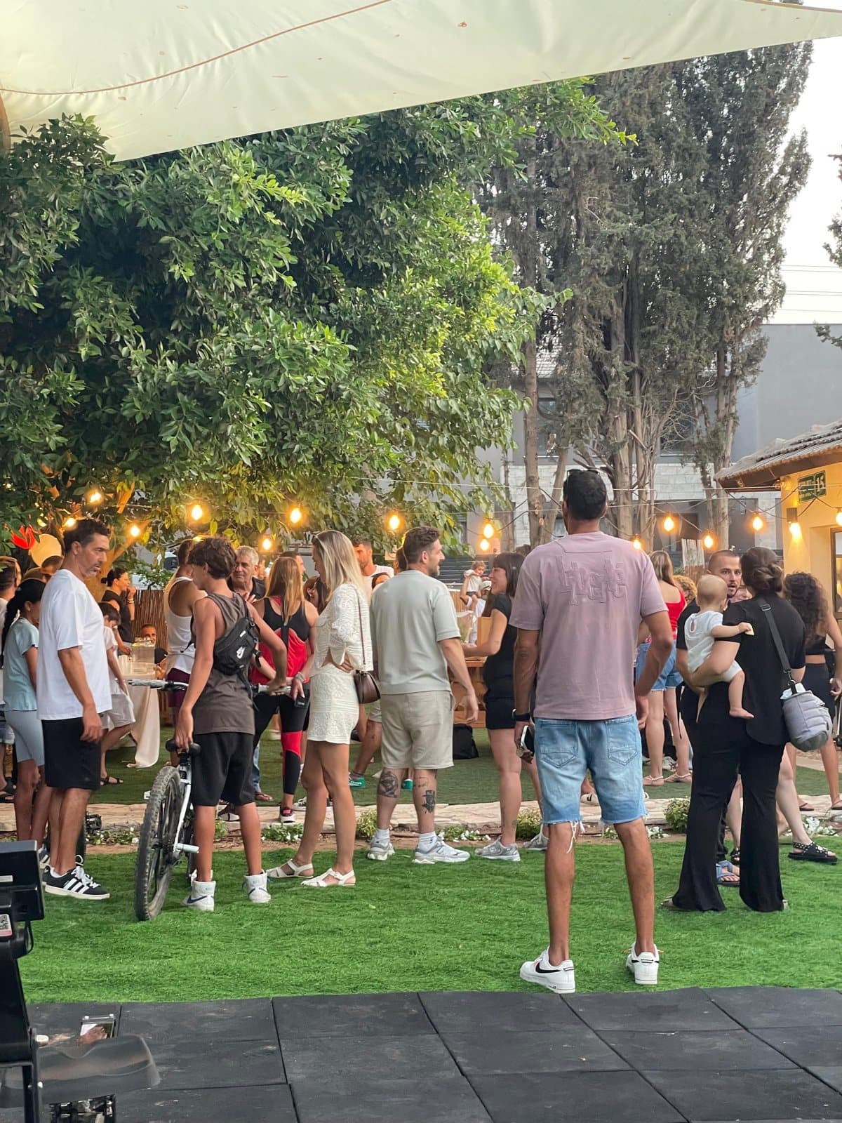 מפגש קהילה תחת הסוכך: קהל גדול עומד על הדשא בשעת ערב