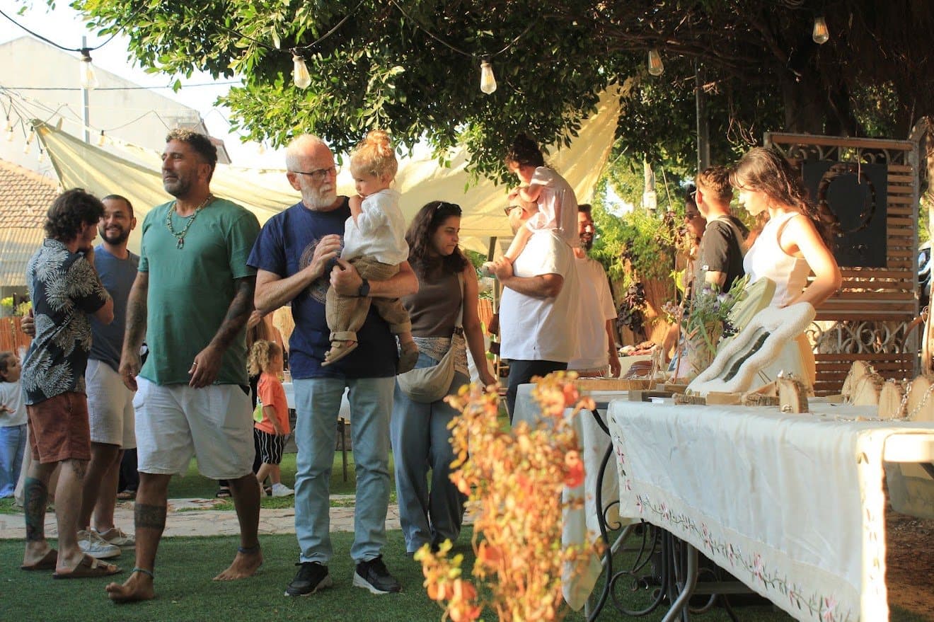 יריד קהילה בחצר: משפחות עוברות בין דוכנים תחת עץ גדול